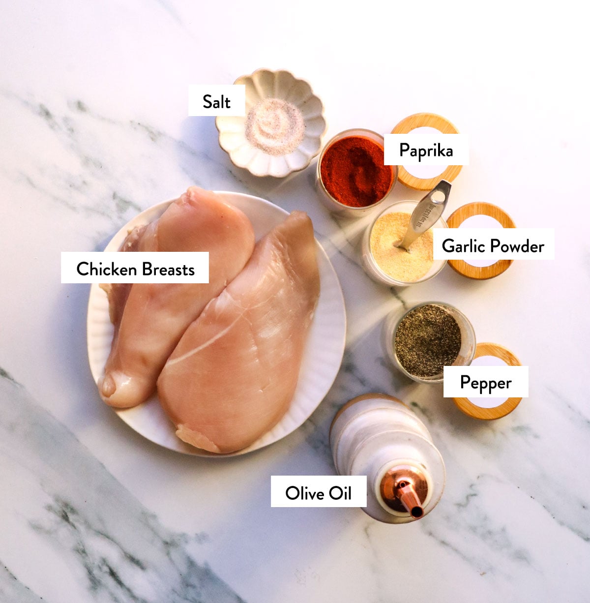 chicken breast, salt, garlic powder, paprika, pepper, and olive oil labeled on a white surface.