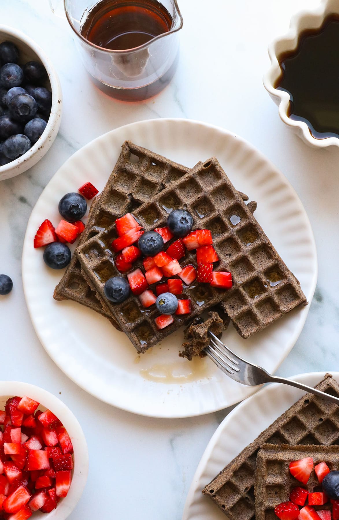 buckwheat waffles on a plate with fruit and maple syrup on top.