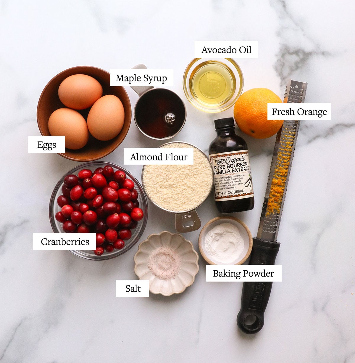almond flour, eggs, maple syrup, cranberries, fresh orange, oil, and vanilla extract labeled on a white surface.