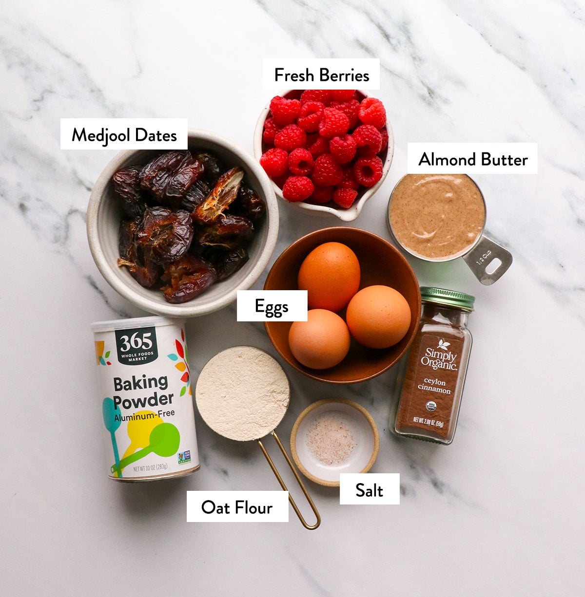 bowls of dates, raspberries, oat flour, eggs, and nut butter labeled on a white surface.