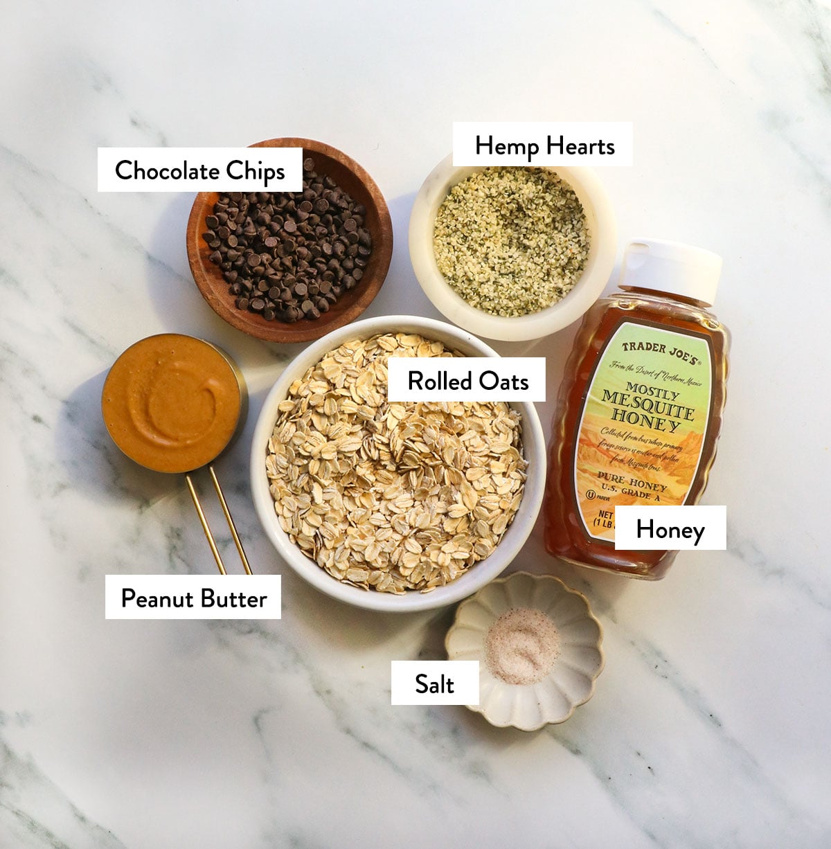 bowl of oats, honey, peanut butter, hemp hearts, and chocolate chips labeled in bowls.