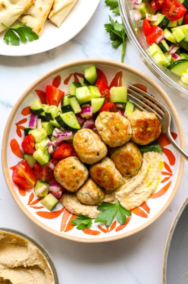Greek chicken meatballs served in a bowl with hummus and cucumber salad.