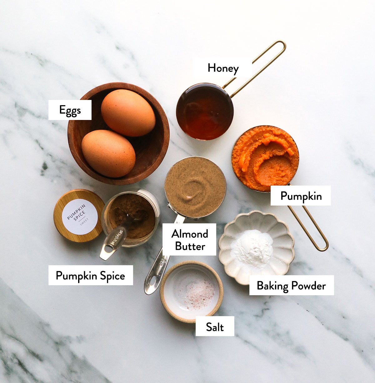 pumpkin puree, almond butter, eggs, honey, and spices labeled on a white surface.