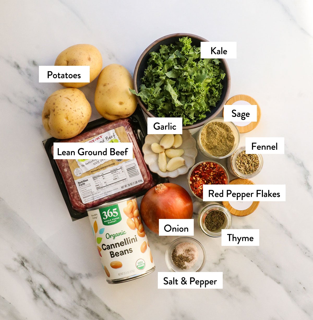 ground beef, potatoes, kale, white beans, garlic, onion, and spices labeled on a white surface.