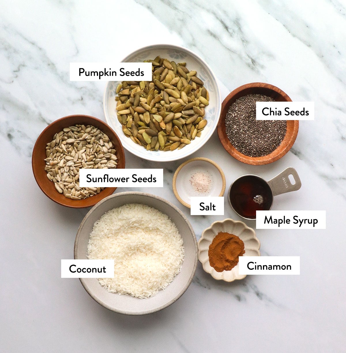 bowls of coconut, pumpkin seeds, chia seeds, maple syrup, and cinnamon labeled.