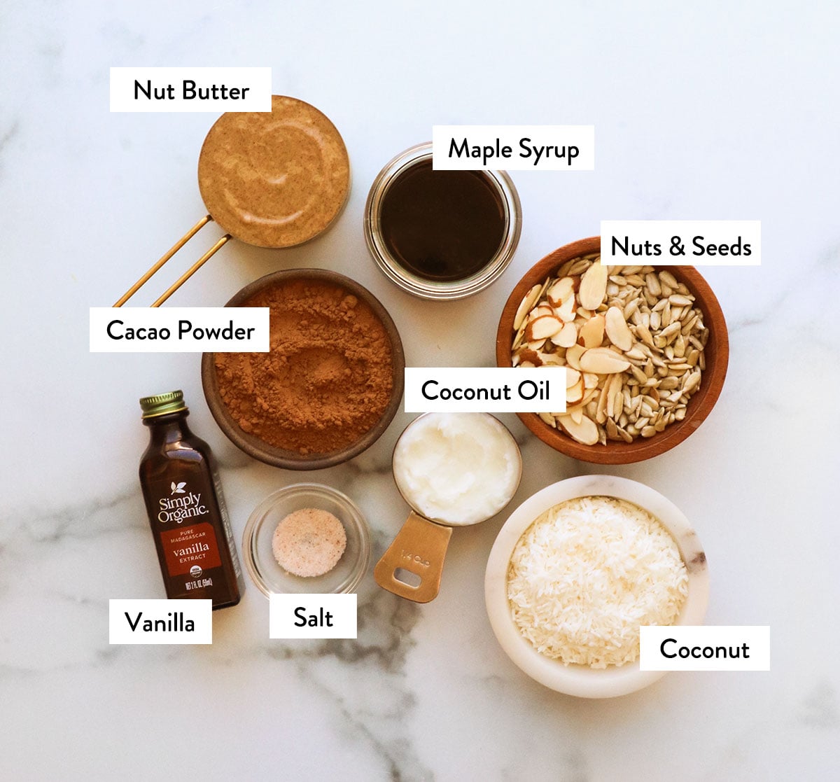 cacao powder, nut butter, coconut, seeds, maple syrup and vanilla labeled on a white surface.